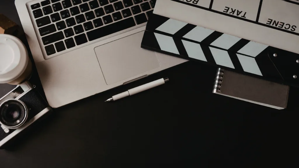 Content creator workspace flatlay with laptop, film clapboard, camera, and notebook on a dark surface.